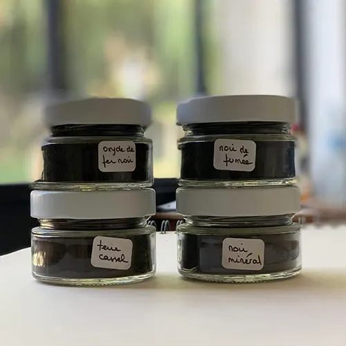 A l'atelier Mylène Mai, devant la verrière, 4 pots de pigments de différentes variations de noirs sont visibles.