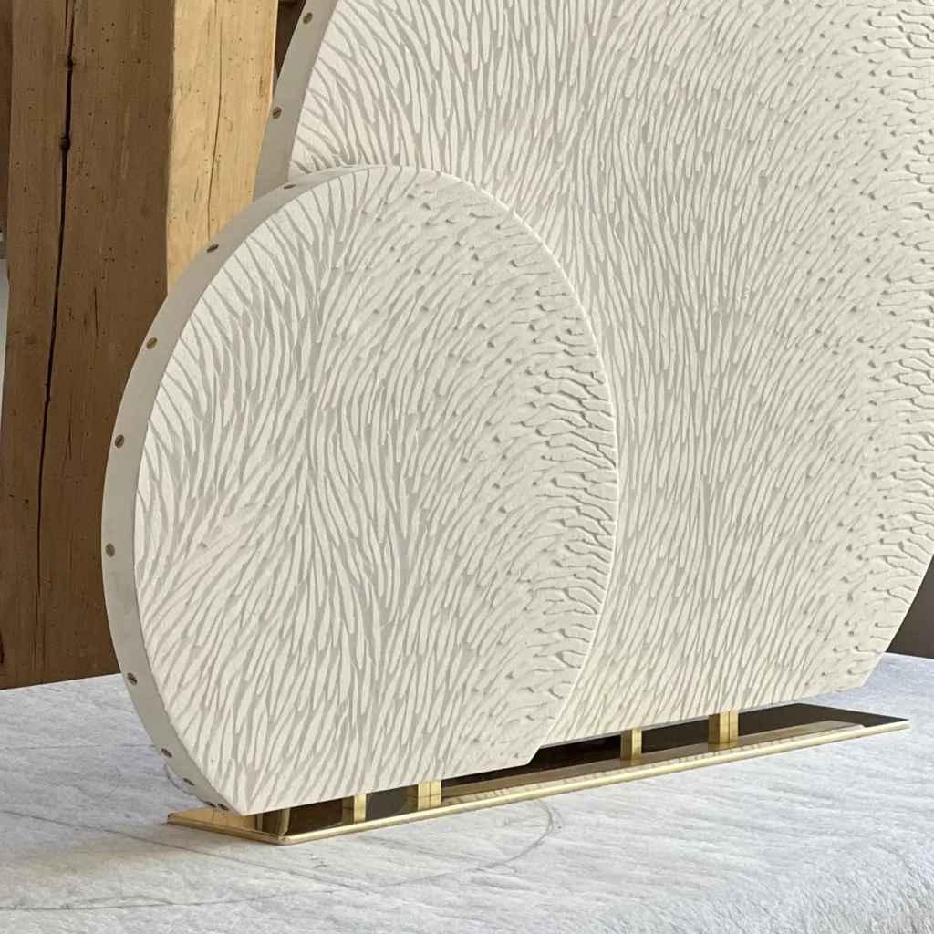 Dans l'atelier Mylène Mai, sculpteur en Bourgogne, on voit une oeuvre en cours, posé sur un plan de travail. IL s'agit d'une sculpture double sur socle, composé de 2 cercles de tailles différentes 40 et 70 cm, tronqués à la base. Les 2 pièces sont sculptés, d'écailles de plâtre brut, pas encore teintées, patinées. En arrière plan, on aperçoit une poutre, ce qui donne un caractère authentique à l'atelier situé dans la campagne Bourguignonne.