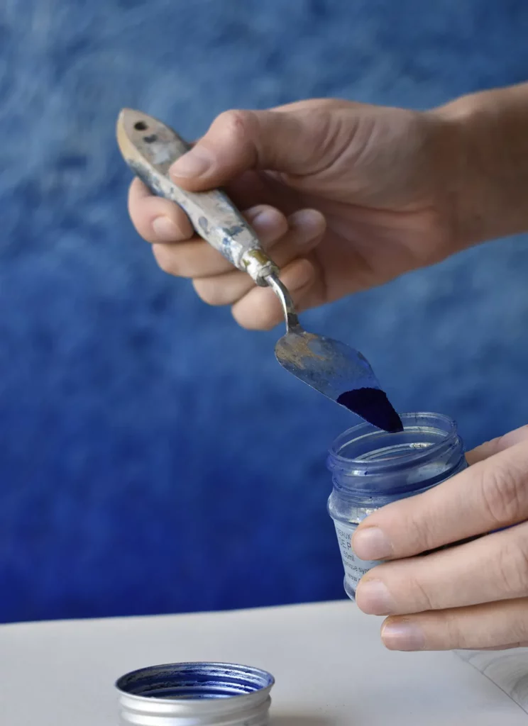 Dans son atelier, les mains de l'artiste Mylène Mai tiennent une spatule et un pot de pigment bleu. En second plan, une œuvre murale sculptée bleu est visible.