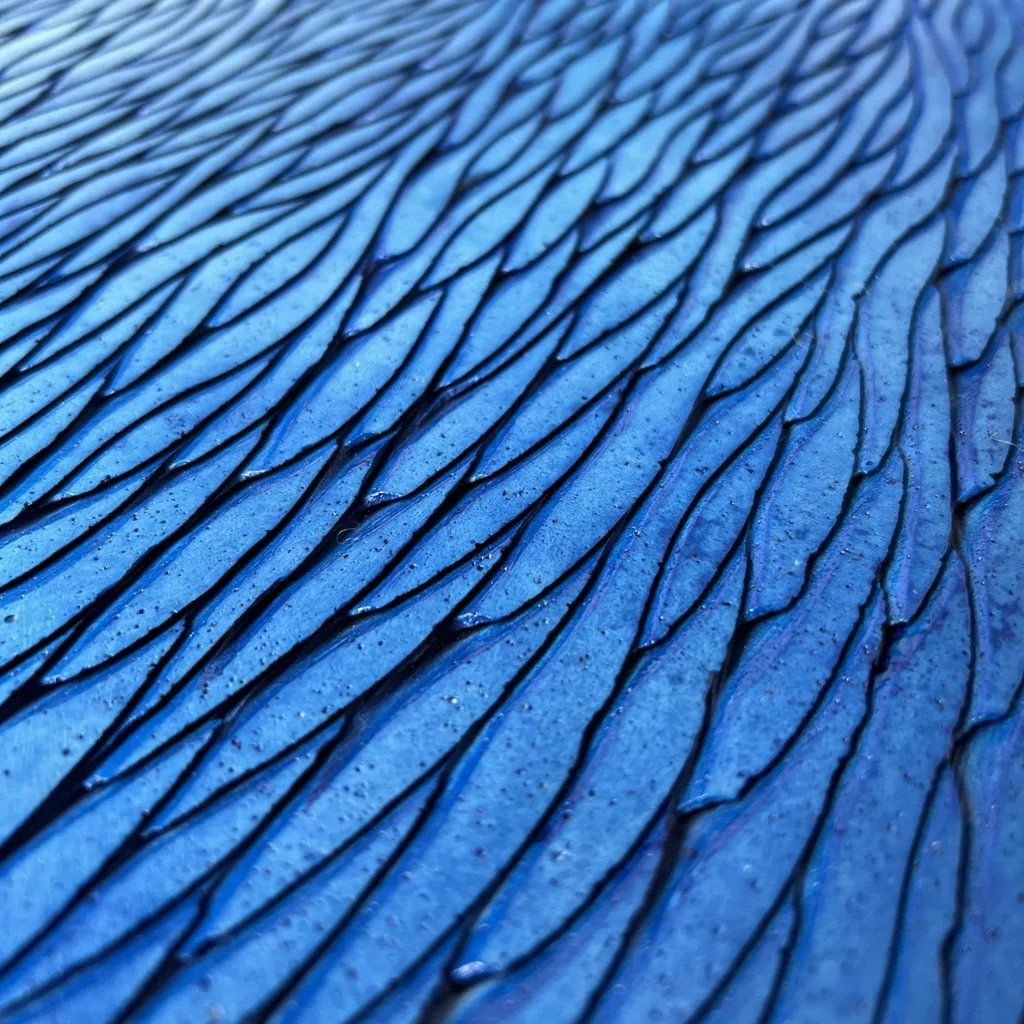 Les écailles de plâtre sculptées et patinées de teintes de bleu sont visibles en zoom, légèrement de trois quart. Les couleurs sont en dégradés, doux et lumineux.