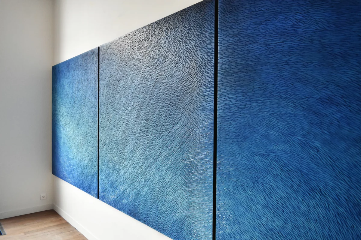 Dans l'atelier de Mylène Mai en Bourgogne, un immense triptyque bleu est accroché sur les murs blancs. On voit de trois quart l'œuvre légèrement zoomée, il s'agit d'une sculpture murale contemporaine, constituée d'une multitudes d'écailles de plâtre sculptées. Cette pièce de très grand format est teintée de nuances de pigments bleu, outremer, azur, en dégradé. L'atelier est baigné de lumière naturelle, réchauffé d'un parquet chêne ancien.