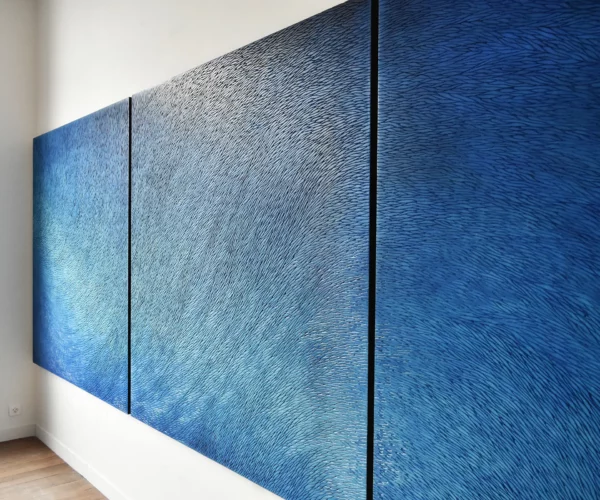 Dans l'atelier de Mylène Mai en Bourgogne, un immense triptyque bleu est accroché sur les murs blancs. On voit de trois quart l'œuvre légèrement zoomée, il s'agit d'une sculpture murale contemporaine, constituée d'une multitudes d'écailles de plâtre sculptées. Cette pièce de très grand format est teintée de nuances de pigments bleu, outremer, azur, en dégradé. L'atelier est baigné de lumière naturelle, réchauffé d'un parquet chêne ancien.