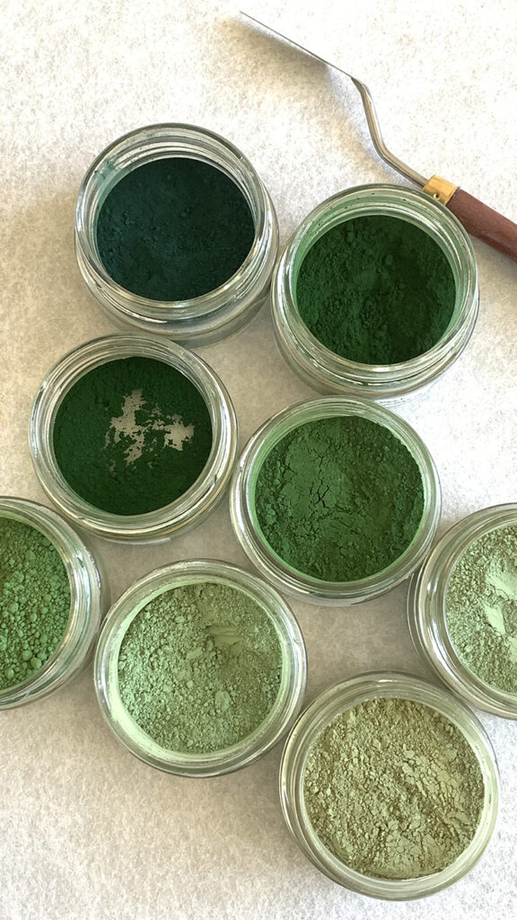 Dans l'atelier de Mylène Mai, une vue de dessus sur des pots de pigments ouverts, posés sur un plan de travail, une spatule posée à côté. Les huit pigments sont tous des teintes de vert différents, du plus clair ou plus foncé et créent une palette de couleur riche.