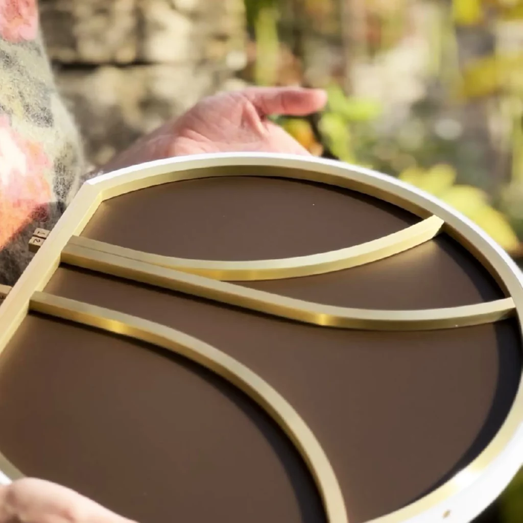 L'artiste Mylène Mai tient dans ses mains, une oeuvre en cours, on voit le dos de la pièce, une strucure en laiton et dos laqué brun. L'œuvre est de forme ronde tronquée de diamètre 40cm.