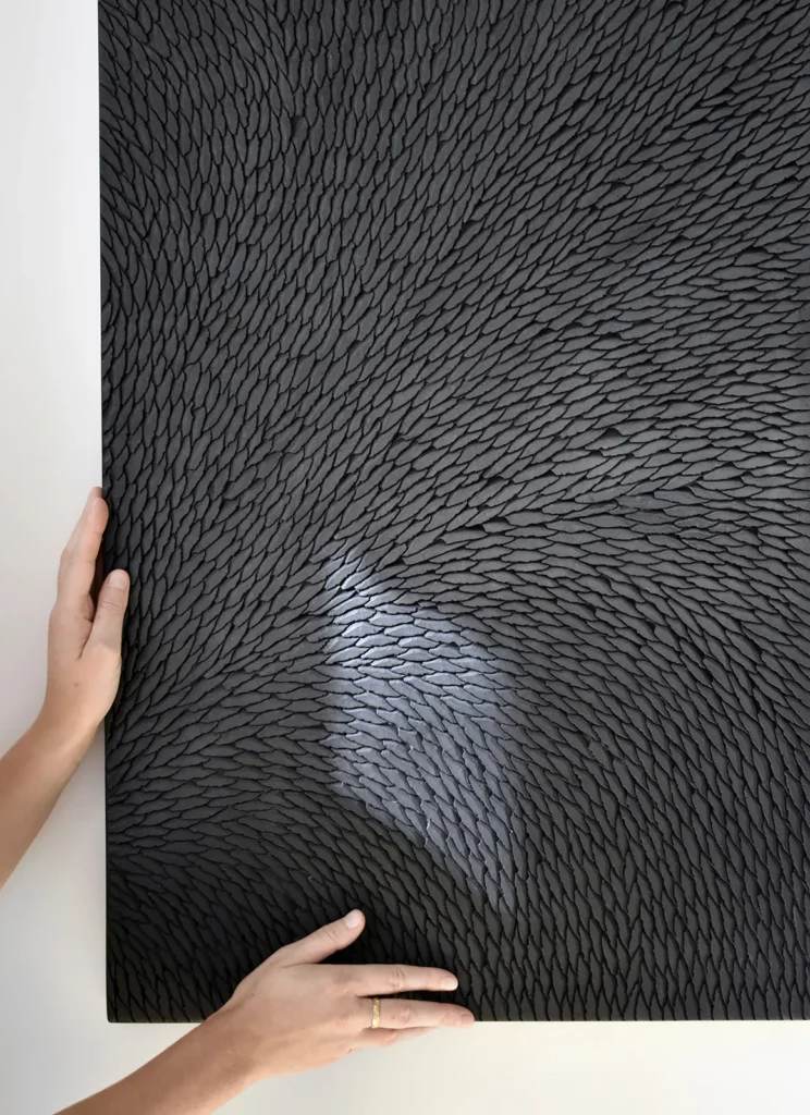 De face, on voit un détail d'une œuvre sculptée, installée au mur blanc. Les mains de l'artiste Mylène Mai finalise l'accrochage de l'oeuvre aux petites écailles de plâtre noir et gris mat.