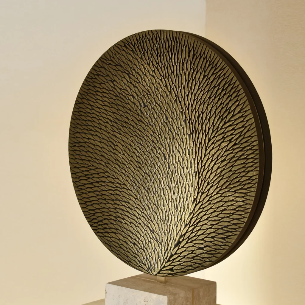 Vue de trois quart d'une sculpture ronde de diamètre cm, sur socle pierre et laiton. La sculpture est en plâtre, composée de multiples petites écailles sculptées et teintées dans des tons de bronze et de doré.