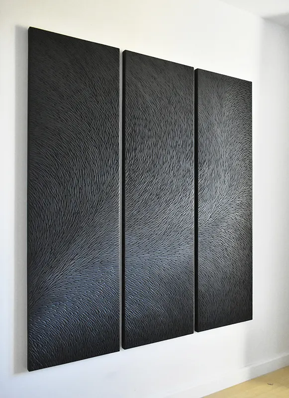 Vue de trois quart d'un triptyque, une sculpture murale de l'atelier Mylène Mai, constitué de multiples petites écailles de plâtre. La patine est réalisée à partir de pigments noir et gris mat, travaillée à l'acrylique et à la cire.