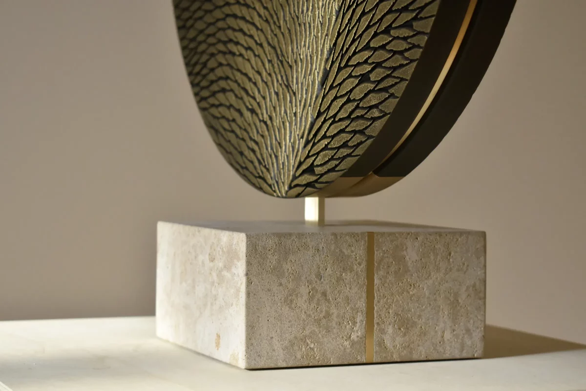 Socle d'une sculpture de l'atelier Mylène Mai, le socle est en pierre de Bourgogne et laiton. Il supporte une oeuvre ronde en laiton et plâtre sculpté. On aperçoit une partie de l'oeuvre en écailles sculptées, petites, fines, parfaitement organisées et teintées de bronze et d'or.