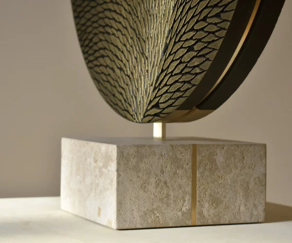 Socle d'une sculpture de l'atelier Mylène Mai, le socle est en pierre de Bourgogne et laiton. Il supporte une oeuvre ronde en laiton et plâtre sculpté. On aperçoit une partie de l'oeuvre en écailles sculptées, petites, fines, parfaitement organisées et teintées de bronze et d'or.