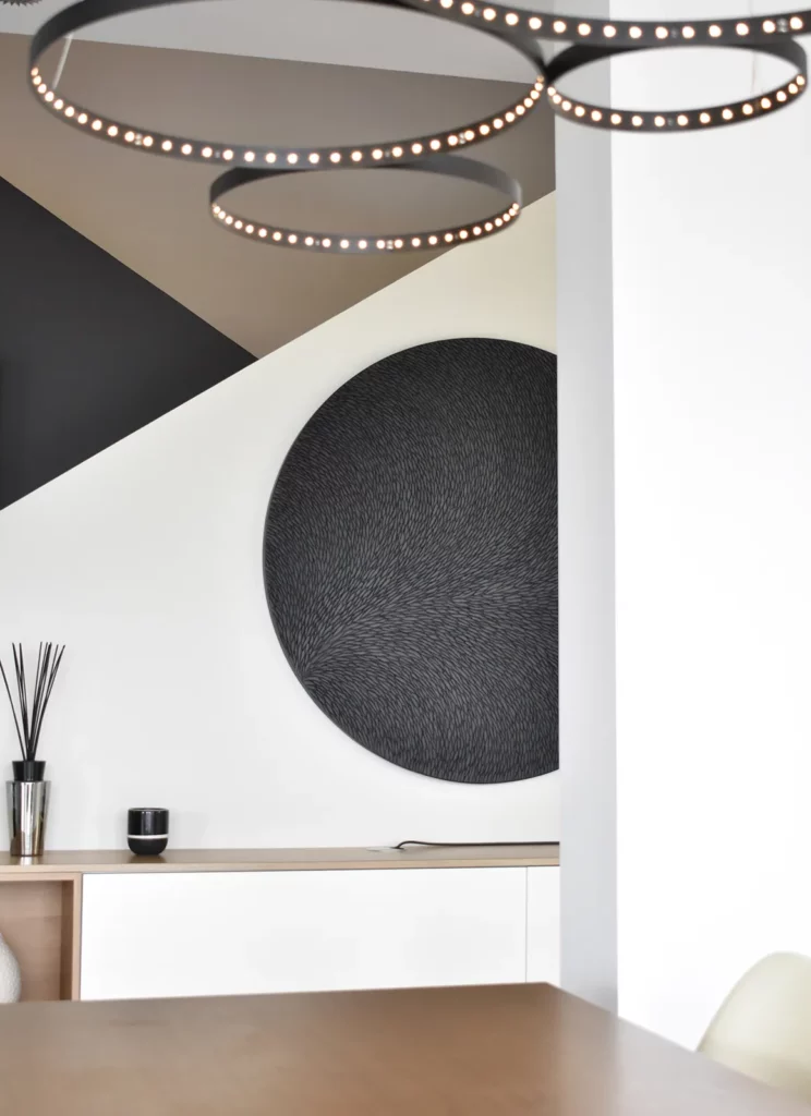 Dans un intérieur contemporain, une sculpture murale ronde de diamètre 100 cm est installée au mur. Mur blanc, luminaire noir et rond en rappel, table à manger en bois, l'intérieur est sobre, épuré.
