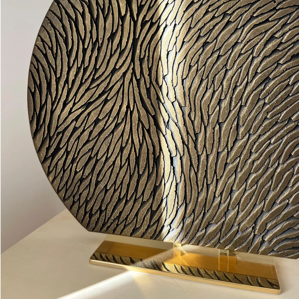 Un rayon de soleil sur une petite sculpture sur socle, à poser sur une étagère, de l'atelier Mylène Mai. L'oeuvre est en écailles de platre sculptée, le socle est en laiton poli miroir. La couleur de l'oeuvre est bronze et or. La forme est un cercle tronqué en bas d'environ 40cm