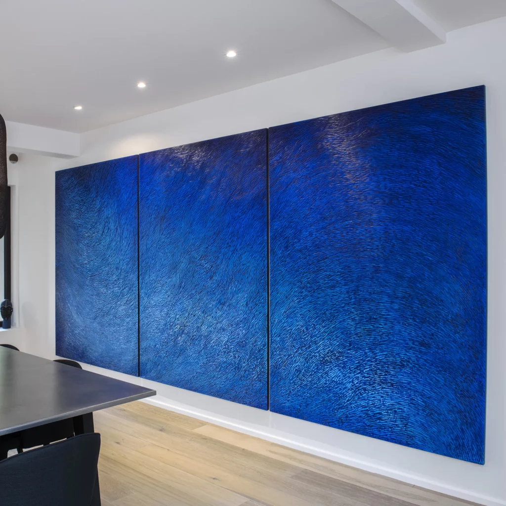 Dans intérieur contemporain, mur blanc, parquet chène, table de designer en métal ... une oeuvre grand format est accroché au mur. C'est un triptyque, une sculpture murale monumentale, d'environ 2 mètres par 4 mètres, entièrement sculpté. Les écailles de plâtre animent la surface, elles sont teintées de multiples pigments bleus, intenses, outremer. Une œuvre contemporaine qui apporte beaucoup de caractère à ce salon moderne.