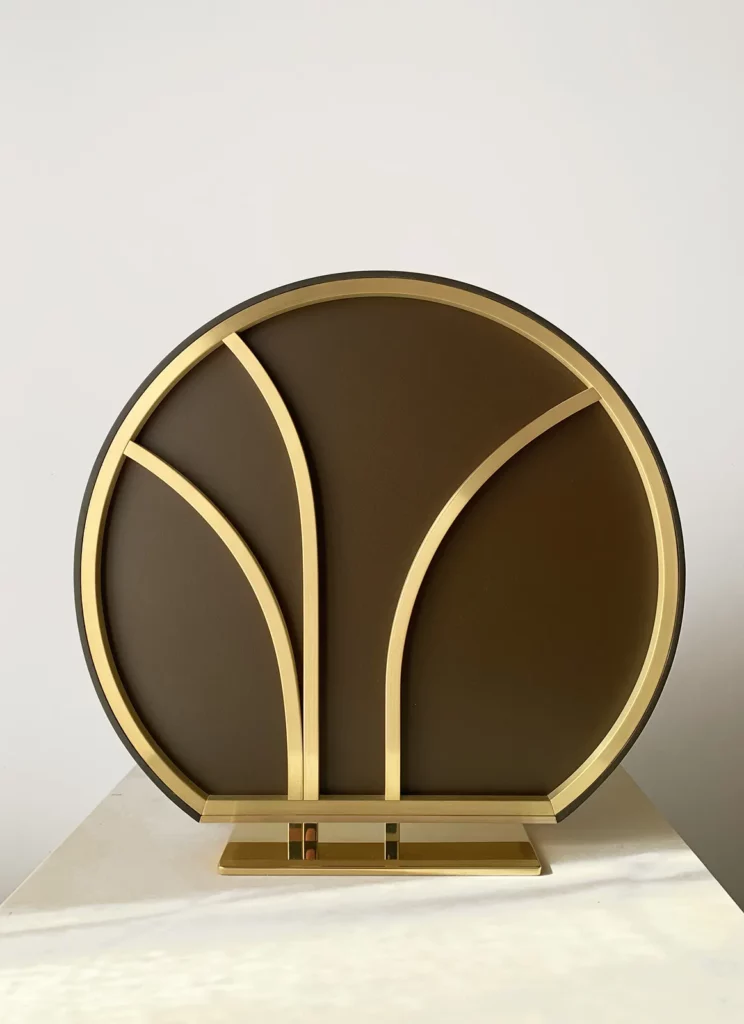 Une sculpture sur socle de l'atelier Mylène Mai, en plâtre et laiton, ici vue de dos, on voit un magnifique travail de structure en laiton, dans un graphisme évoquant l'art déco, un fond laqué brun et un socle en laiton poli miroir. Un bel éventail des savoir-faire et compétences des métiers d'art de l'atelier de sculpture bourguignon Mylène Mai