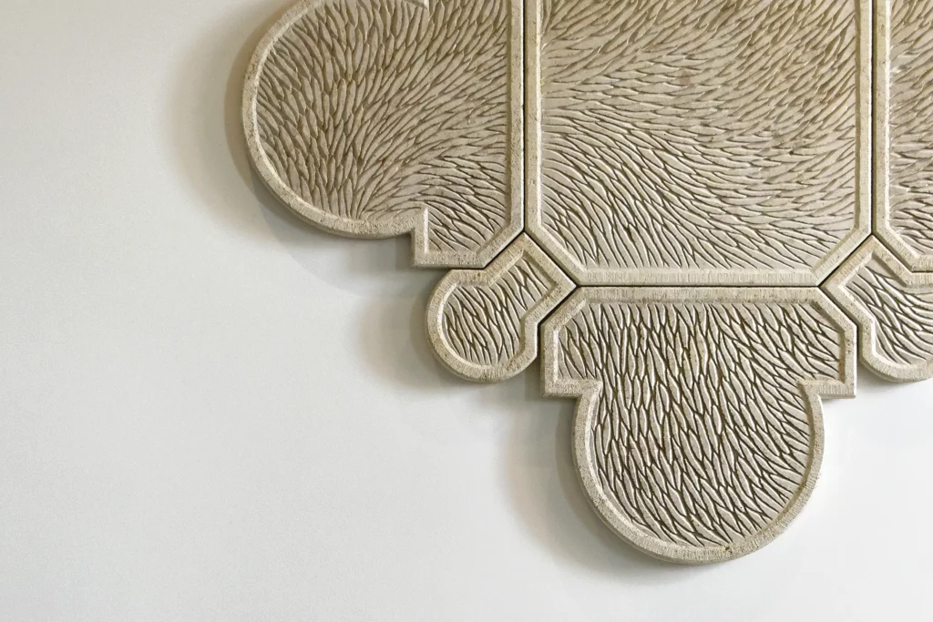 Vue de face partielle d'une œuvre murale sculptée en pierre de Bourgogne. L'oeuvre est composée de plusieurs éléments qui s'assemblent, la forme générale est un motif inspiré de l'architecture, de l'univers floral et de la joaillerie, un contour tout en courbure, symétrique et précis. Toute la surface de l'oeuvre est sculptée d'écailles finement ciselée et organisée selon un mouvement général. La pierre de Bourgogne beige claire est lumineuse, sur un mur blanc type galerie d'art