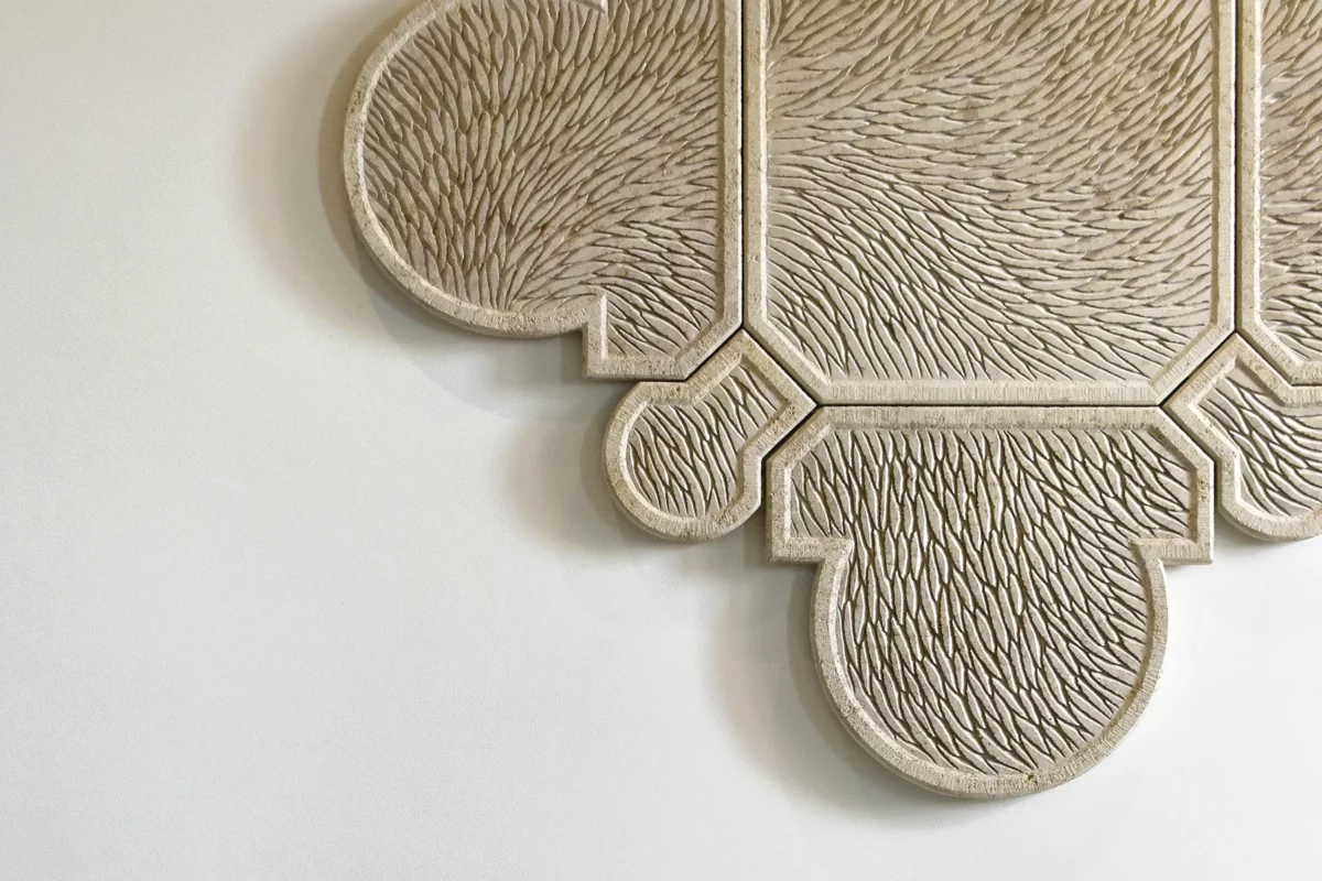 Vue de face partielle d'une œuvre murale sculptée en pierre de Bourgogne. L'oeuvre est composée de plusieurs éléments qui s'assemblent, la forme générale est un motif inspiré de l'architecture, de l'univers floral et de la joaillerie, un contour tout en courbure, symétrique et précis. Toute la surface de l'oeuvre est sculptée d'écailles finement ciselée et organisée selon un mouvement général. La pierre de Bourgogne beige claire est lumineuse, sur un mur blanc type galerie d'art