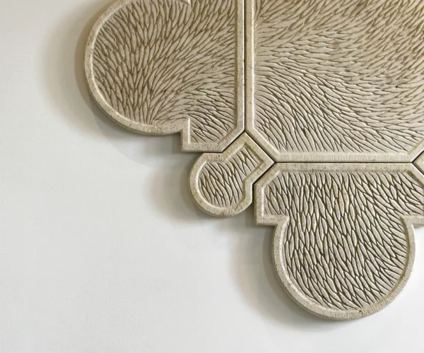 Vue de face partielle d'une œuvre murale sculptée en pierre de Bourgogne. L'oeuvre est composée de plusieurs éléments qui s'assemblent, la forme générale est un motif inspiré de l'architecture, de l'univers floral et de la joaillerie, un contour tout en courbure, symétrique et précis. Toute la surface de l'oeuvre est sculptée d'écailles finement ciselée et organisée selon un mouvement général. La pierre de Bourgogne beige claire est lumineuse, sur un mur blanc type galerie d'art