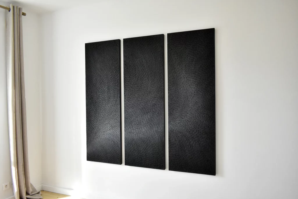 Dans un intérieur aux tons neutres, murs blancs et rideaux beige, une oeuvre de l'artiste Mylène Mai, un triptyque noir et gris mat, est accroché au mur, visible de trois quart. Les écailles de plâtre sculptées captent la lumière.