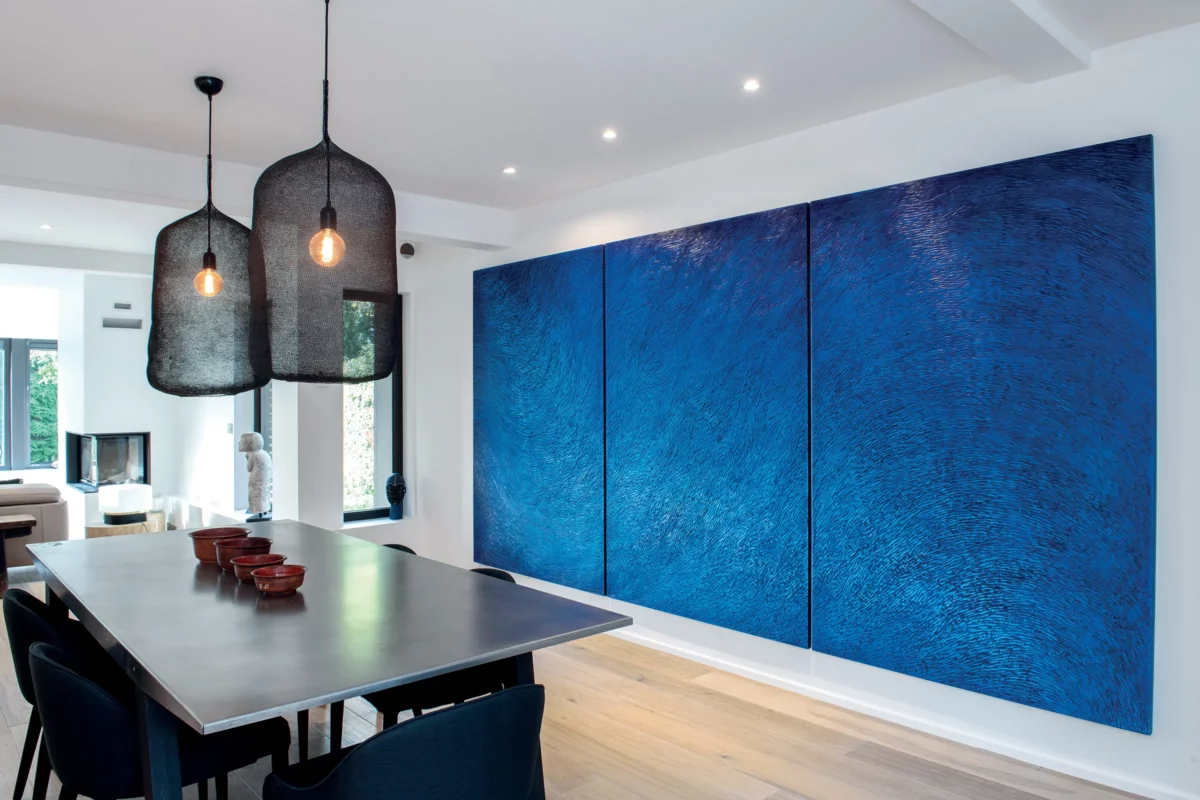 Une maison d'architecte contemporaine, avec un mobilier minimaliste en métal noir, des suspensions noires, huisseries noires, un parquet chêne, des murs blancs. Une oeuvre d'art forte et imposante est accrochée au mur, un triptyque bleu outremer, une sculpture murale de l'atelier Mylène Mai.