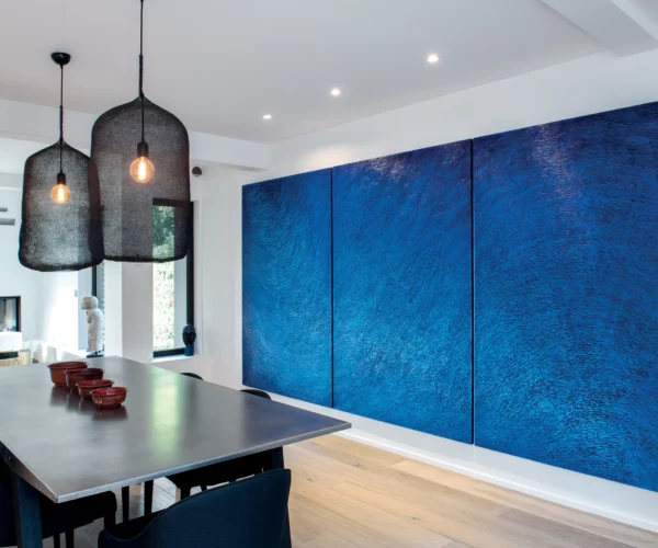 Une maison d'architecte contemporaine, avec un mobilier minimaliste en métal noir, des suspensions noires, huisseries noires, un parquet chêne, des murs blancs. Une oeuvre d'art forte et imposante est accrochée au mur, un triptyque bleu outremer, une sculpture murale de l'atelier Mylène Mai.