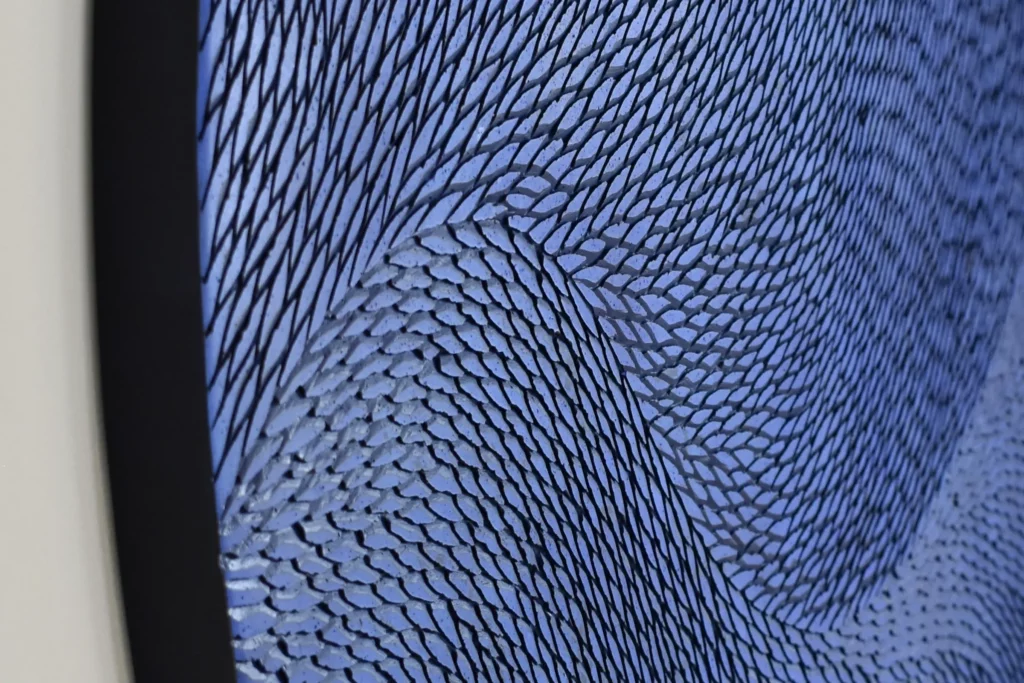 Un zoom sur une oeuvre murale en petites écailles de plâtre sculptées. L'oeuvre est installée sur un mur blanc type galerie d'art. Les petites écailles sont teintées de pigments bleu intenses de type outremer.