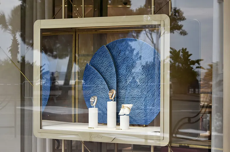 Vue extérieure d'une vitrine de la maison Cartier à Cannes, légèrement de trois quart. On voit une sculpture sur socle de l'atelier Mylène Mai, une forme ronde composée de 4 pièces en volume, sculptées d'écailles de plâtre, teintées de nuances de bleus. C'est un décor et un écrin à la joaillerie présentée.