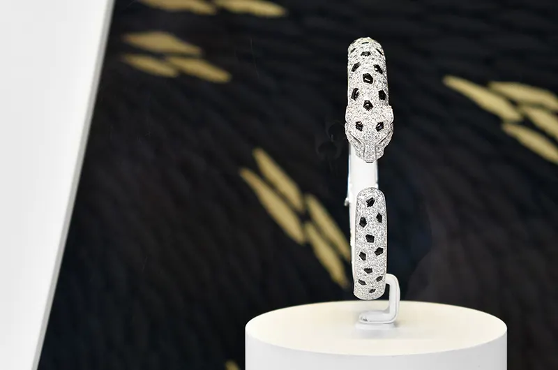 On aperçoit au premier plan, un bracelet de haute joaillerie de la maison Cartier, une collection panthère. En arrière plan, on reconnaît le travail de l'artiste Mylène Mai, par ces écailles de plâtre sculptées, ici teintées de noir et quelques une dorées pour évoquer le pelage de l'animal emblématique de la maison Cartier, client et commanditaire de cette œuvre d'exception sur mesure.