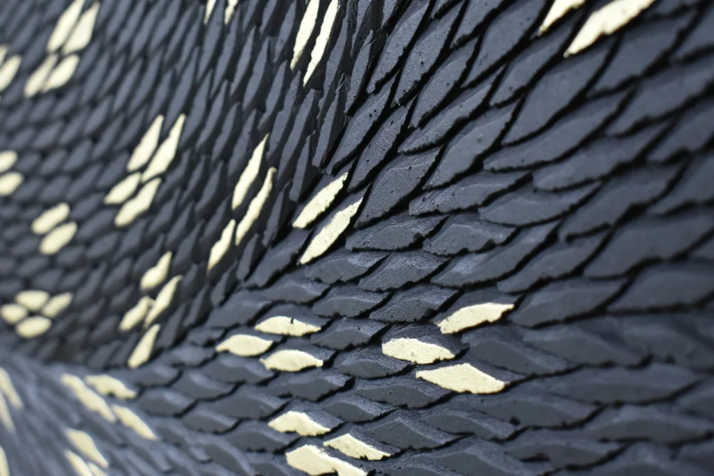 Zoom sur la matière d'une oeuvre de l'atelier Mylène Mai. L'oeuvre sur mesure est une pièce unique constitué de multiples petites écailles de plâtre sculpté. La teinte de l'ensemble est noir mat, avec quelques écailles dorées à la main, regroupés par petits groupes de 3 ou 4, pour évoquer le pelage de la panthère, animal emblématique de la maison Cartier, client et commanditaire de cette oeuvre d'exception.