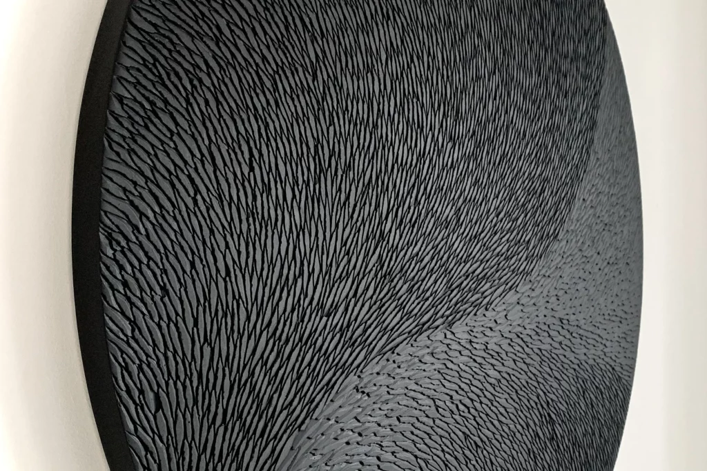 Vue zoomée et de trois quart sur une sculpture murale ronde de l'artiste Mylène Mai. L'oeuvre est constituée de multiples petites écailles de plâtre sculptées, animée par un mouvement végétal et organique parfaitement orchestré. La teinte de l'oeuvre est un subtil dégradé de noir et de gris mat. L'oeuvre est fine, environ 3 cm d'épaisseur.