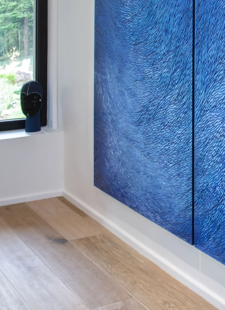 C'est un détail vu de trois quart d'une oeuvre murale, une sculpture monumentale en plâtre, installée chez les clients, dans une maison contemporaine, une villa d'architecte aux huisseries modernes, murs blancs et parquet chêne. L'œuvre en plâtre sculptée, composées d'écailles, est teintée de différents pigments bleus, cela crée des dégradés de couleurs, très lumineux