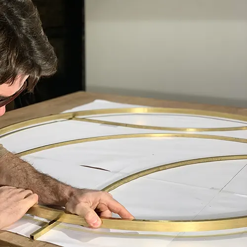 A l'atelier Mylène Mai, on aperçoit Julien qui cintre le laiton et ajuste l'assemblage d'une structure ronde, qui servira de support pour créer une sculpture murale en plâtre. La structure est posée sur un plan à l'échelle, disposé sur le plan de travail.