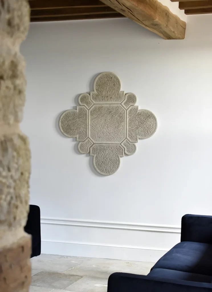 Dans un salon de maison de caractère, avec poutres en chêne, sol en pierre, les murs blancs avec fine moulure au dessus de la plinthe sont orné d'une sculpture murale fine en pierre de Bourgogne. La forme est complexe, composée de 9 éléments qui composent un motif inspiré de l'architecture. Dans ce salon, 2 canapés bleus nuit se font face devant l'oeuvre. L'ensemble est très épuré, mêlant contemporain et caractère historique.