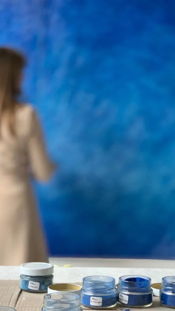 Au premier plan, des pots de différents pigments bleus sont ouverts, posés sur un plan de travail à l'atelier en Bourgogne. Au second plan, légèrement flouté, Mylène Mai travaille à la réalisation de la patine sur un grand format monumental.