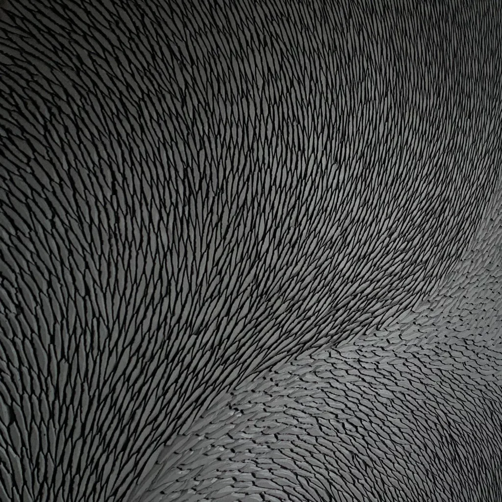 Zoom sur la matière sculptée par l'atelier Mylène Mai, ici une oeuvre en plâtre constituée de multiples toutes petites écailles, fines et parfaitement organisées. L'oeuvre est teintée de pigments noir et gris mat.