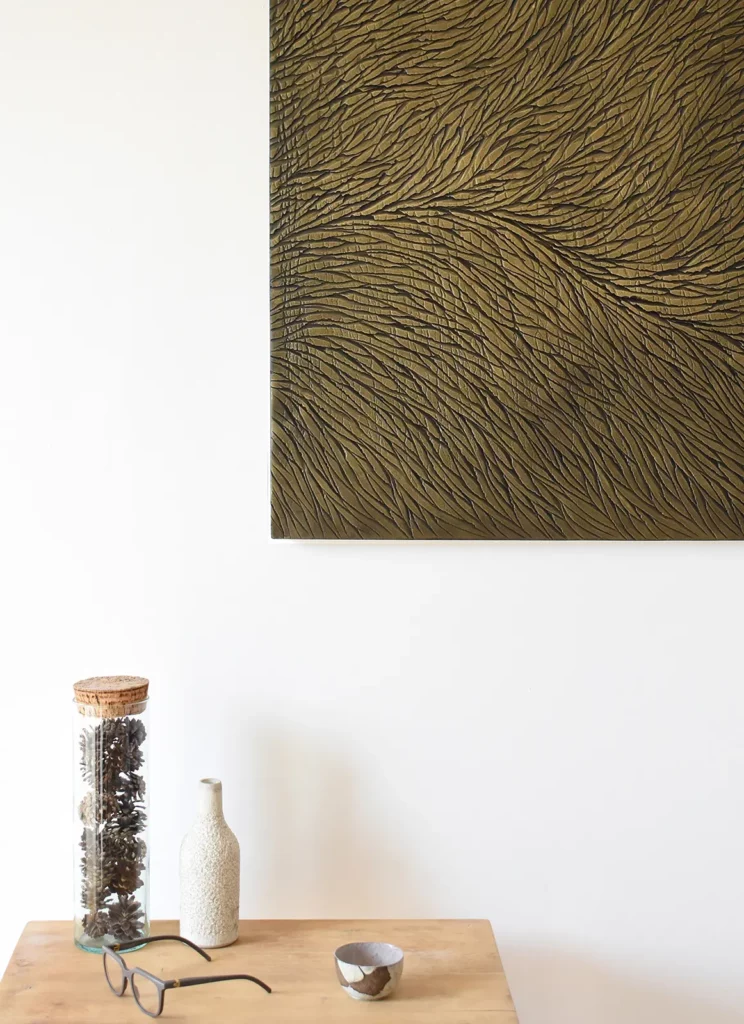 Une sculpture de l'atelier Mylène Mai, vue partiellement, exposée dans un intérieur mur blanc type galerie, table basse avec décoration, lunettes de l'artiste, une tasse de thé. L'ensemble dans des tons neutres, bois, pierre, esprit wabi sabi.