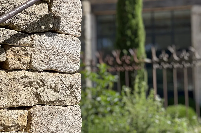 Au premier plan, un mur de jardin en pierre de Bourgogne et une rambarde ancienne en fer forgé, plus loin, un massif avec un cyprès et la grande verrière de l'entrée de l'atelier Mylène Mai