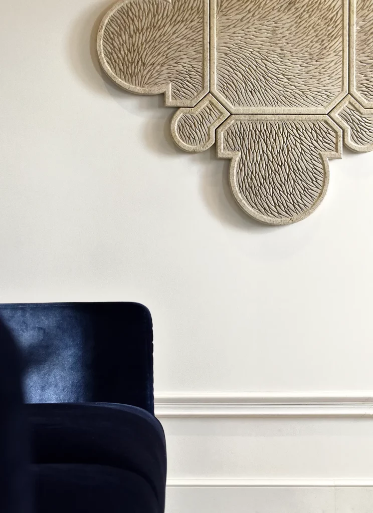 Dans un salon aux murs blancs mouluré, à gauche un canapé en velours bleu partiellement visible, au mur une partie d'une oeuvre murale sculptée en pierre de Bourgogne, une forme inspirée de l'architecture, d'un motif floral ou issu de la joaillerie. La composition est épurée, à la fois moderne et historique.