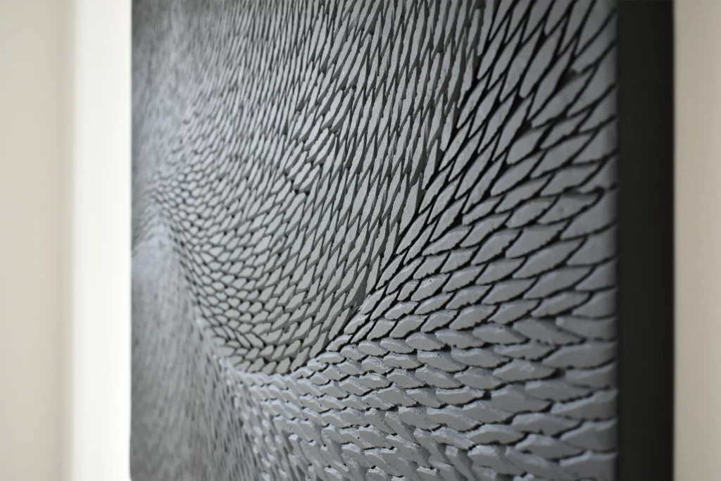 Vue de profil d'une oeuvre de l'artiste Mylène Mai, composée de multiples petites écailles de plâtre noir et gris mat. Le mouvement des écailles captent la lumière et créent une sensation de mouvement.