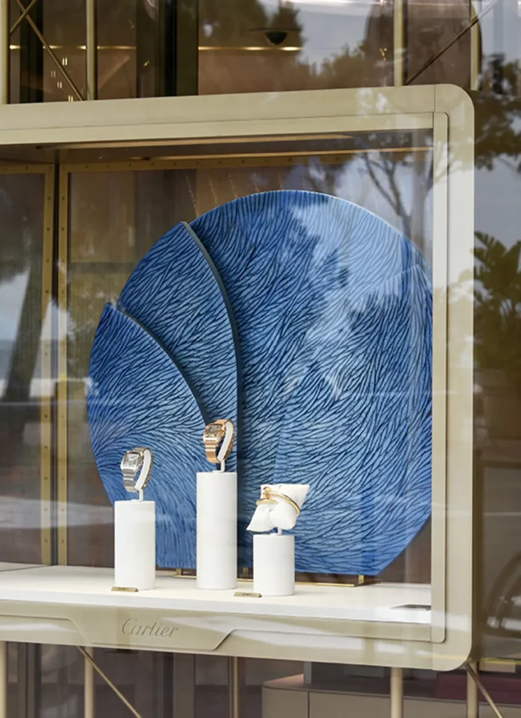 Vitrine vue depuis l'extérieur de la boutique Cartier Cannes, une oeuvre sur mesure, pièce unique réalisée par l'atelier Mylène Mai est disposée comme un décor ou un écrin à deux créations horlogères et un bracelet de la maison Cartier. L'oeuvre sur socle est de forme ronde tronquée à la base est constituée de 4 pièces qui forme le cercle. Elle est entièrement sculptée d'écailles de plâtre, patinées de multiples teintes de bleu dans les tons azur.