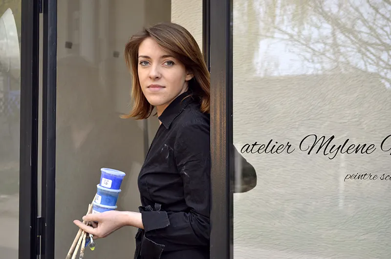 L'artiste sculpteur Mylène Mai sortant de la verrière de son atelier en Bourgogne avec des pigments et des pinceaux à la main.