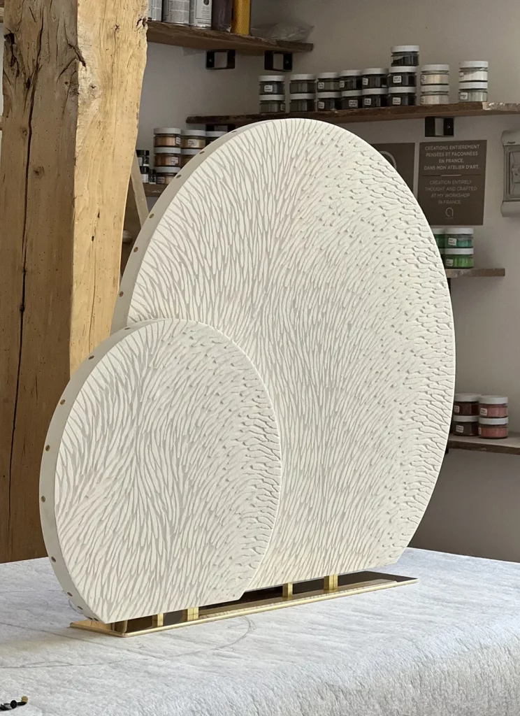 Dans l'atelier de Mylène Mai en Bourgogne, sur un plan de travail est disposée une sculpture double, en cours de réalisation, composée de 2 cercles de diamètre différents 40 et 70 cm, tronqués à la base. Les 2 pièces sont en plâtre sculpté, encore brut, assemblées sur un socle en laiton poli miroir