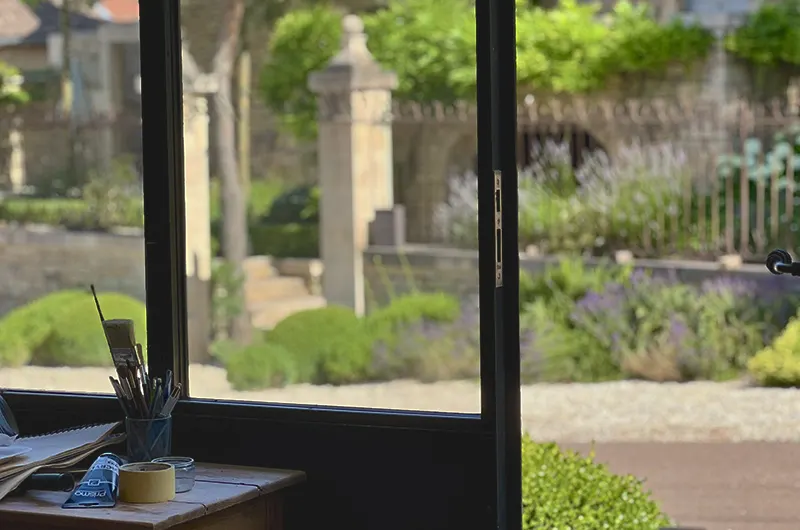 Vue depuis l'intérieur de l'atelier, on aperçoit un petit plan de travail devant la verrière, avec quelques outils, la vue est face à une belle maison ancienne en pierre de Bourgogne, avec beaucoup de charme et d'authenticité, avec un joli jardin historique devant, parfaitement entretenu