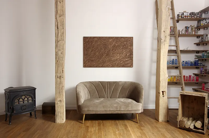 intérieur de l'atelier Mylène Mai en Bourgogne, parquet chêne, canapé beige, poêle à bois, étagère remplie de pigments et sculpture murale bronze doré au mur.
