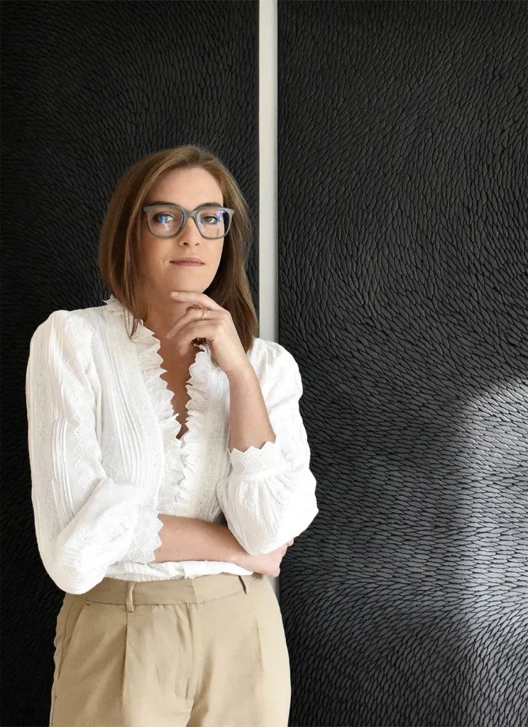 Portrait de l'artiste Mylène Mai, devant un triptyque mural en plâtre sculpté, fait d'une multitude de petites écailles patinées de noir et de gris mat.